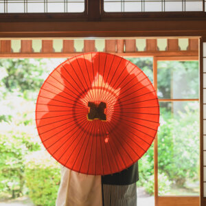 東山荘にて、障子の前で赤い和傘に隠れる新郎新婦の足元だけが見える一枚。和傘を使った和装前撮りの遊び心のあるカット