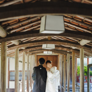 木の梁の連なる渡り廊下で寄り添う黒紋付袴の新郎と白無垢の花嫁。日本庭園の渡り廊下で撮る和装前撮り