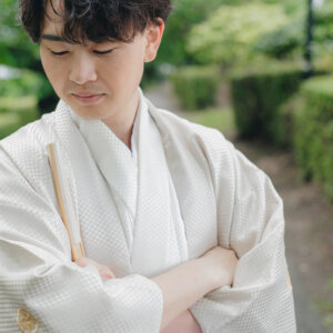 末広を手に腕を組み、目を伏せる白い羽織袴の新郎の上半身カット。和装前撮りの新郎ポートレート
