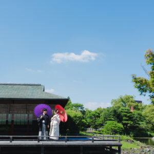 青空の下、蓮池に面したデッキで紫と赤の和傘を差す新郎新婦を引きでとらえた一枚。白鳥庭園で撮る和装ロケーションフォト