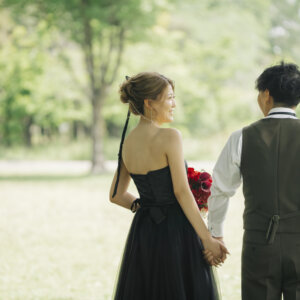 新緑の公園で手をつないで歩くオリーブのベストの新郎と赤いブーケを持つ黒いドレスの花嫁の後ろ姿。公園で撮る洋装前撮り