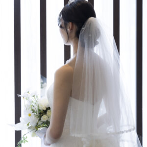ロングベールをまとった花嫁が窓辺に立つ後ろ姿。ティアラとまとめ髪が映えるバックスタイルのウェディングフォト