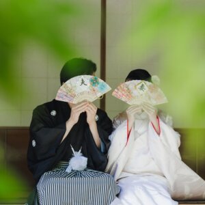 『夫』『妻』の扇子で顔を隠す黒紋付袴の新郎と白無垢の花嫁を、緑の葉越しにとらえた一枚。名古屋のスタジオで撮る和装前撮りの扇子プロップス