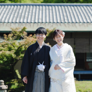 瓦屋根の日本家屋と石灯籠を背に並ぶ黒紋付袴の新郎と白無垢の花嫁。日本庭園で撮る和装前撮り