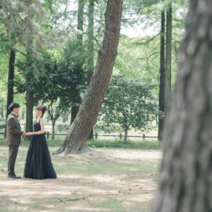 松の木立の中で手を取り合うオリーブのスーツの新郎と黒いドレスの花嫁を引きでとらえた一枚。公園で撮る洋装前撮り