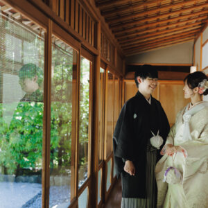 ガラス戸の廊下で向かい合う黒紋付袴の新郎とブーケを持つ白い色打掛の花嫁。日本家屋で撮る和装前撮り