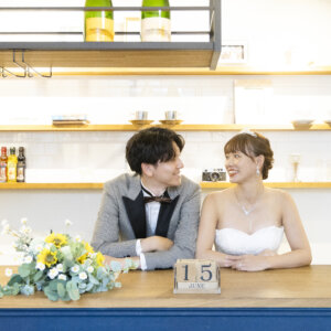 キッチンカウンターに並んで座り、ひまわりのブーケと日付ブロックを飾って笑う新郎新婦。カフェ風スタジオで撮るウェディングフォト
