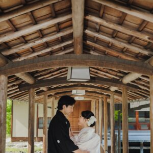 木の梁の連なる渡り廊下で抱き合う黒紋付袴の新郎と白無垢の花嫁を縦位置でとらえた一枚。白鳥庭園で撮る和装前撮り
