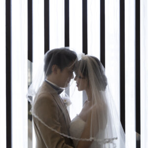 花嫁のベールの中でおでこを寄せ合う新郎新婦。縦格子の窓からの光に包まれたロマンチックな前撮り写真