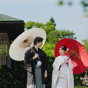 簾の下がる日本家屋の前で白と赤の和傘を差す黒紋付袴の新郎と白無垢の花嫁。和傘を使った和装前撮り