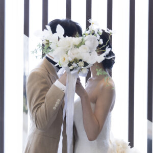 大きな白いブーケで顔を隠してキスする新郎新婦。光が差し込む縦格子の窓辺で撮る、顔隠しポーズのフォトウェディング