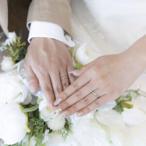 白いバラのブーケの上に手を重ね、結婚指輪を見せる新郎新婦の手元をとらえたウェディングフォトのディテールカット