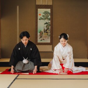 掛け軸の前で赤い毛氈に正座して手をつく黒紋付袴の新郎と白無垢の花嫁。和室で撮る和装前撮りのお辞儀ポーズ