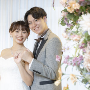 花の装飾の前で頬を寄せ、手を重ねて微笑むグレーのタキシードの新郎とティアラの花嫁。名古屋のスタジオで撮る洋装前撮り