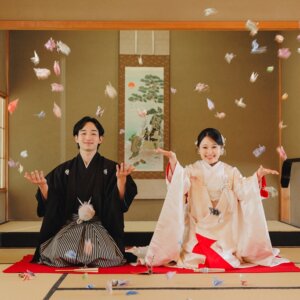 白鳥庭園にて、和室で折り鶴を投げ上げて笑う黒紋付袴の新郎と白無垢の花嫁。折り鶴を使った和装前撮りのアイテムカット