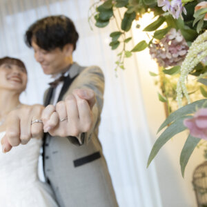 花の壁の前で、結婚指輪をはめた手をこぶしにしてカメラへ差し出す新郎新婦。指輪を主役にしたウェディングフォトのポーズ