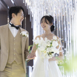 手前の装花をぼかし、手を取り合って笑い合う新郎と白いブーケを持つ花嫁を捉えたナチュラルなウェディングフォト