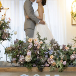 花の装飾とイニシャルオブジェの奥で、抱き合っておでこを寄せる新郎新婦。前ボケを生かしたスタジオフォトウェディング