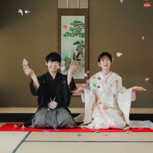 和室で折り鶴を投げ上げて笑う黒紋付袴の新郎と白い色打掛の花嫁。折り鶴を使った和装前撮りのアイテムカット