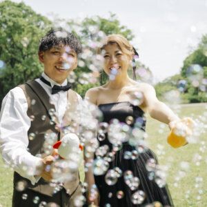 庄内緑地にて、芝生の公園でシャボン玉を飛ばして笑うオリーブのベストの新郎と黒いドレスの花嫁。シャボン玉を使った洋装前撮り
