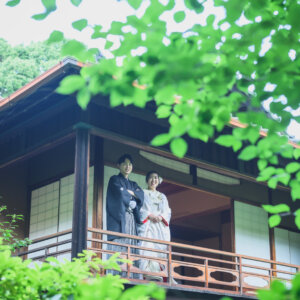 新緑の葉越しに、2階の縁側に立つ黒紋付袴の新郎と白無垢の花嫁をとらえた一枚。東山荘で撮る和装ロケーションフォト