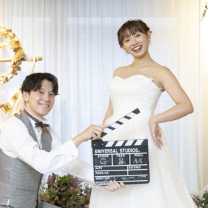 ひざまずいてカチンコを差し出す新郎と、腰に手を当てて笑うウェディングドレスの花嫁。花の装飾の前で撮る遊び心のあるウェディングフォト