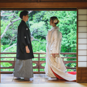 障子の枠越しに、縁側で向かい合う黒紋付袴の新郎と白無垢の花嫁をとらえた一枚。日本家屋で撮る和装前撮り