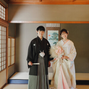 掛け軸の前でピンクのブーケを持って立つ白い色打掛の花嫁と黒紋付袴の新郎を縦位置でとらえた一枚。和室で撮る和装前撮り