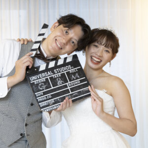 カチンコを手に顔を寄せて笑う、グレーのベストの新郎とウェディングドレスの花嫁。映画のワンシーン風のプロップスを使ったスタジオ前撮り