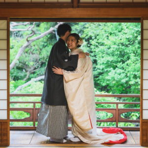 障子の枠越しに、縁側で抱き合う黒紋付袴の新郎と白無垢の花嫁をとらえた一枚。東山荘で撮る和装前撮り