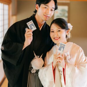 和室でチェキの写真を持って笑う黒紋付袴の新郎とピンクの色打掛の花嫁。インスタントカメラを使った和装前撮りのアイテムカット