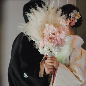 ピンクのパンパスグラスのブーケで顔を隠す白い色打掛の花嫁と黒紋付袴の新郎。ブーケを使った和装前撮りの遊び心のあるカット
