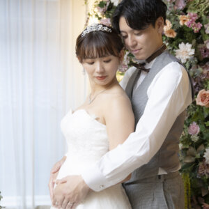 花の壁の前で、後ろから花嫁のお腹に手を添えてバックハグする新郎。ティアラとウェディングドレスのやさしい雰囲気のスタジオ前撮り