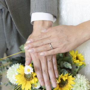 ひまわりのブーケの上に重ねた新郎新婦の手元のアップ。結婚指輪が並ぶウェディングフォトのディテールカット