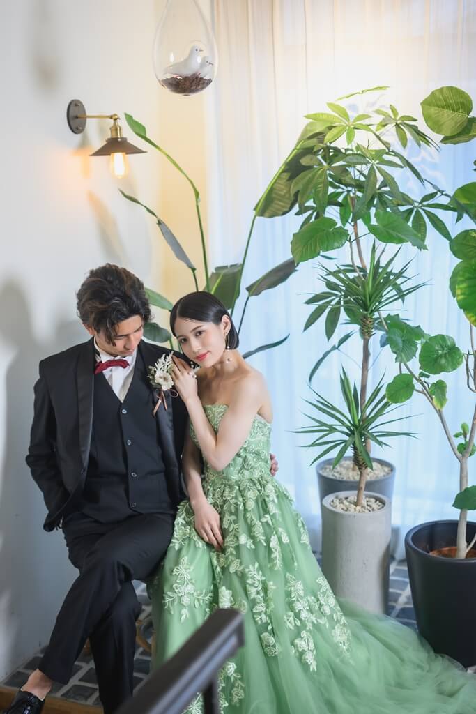 レトロなブラケットライトの下で寄り添って座る、グリーンのレースドレスの花嫁と黒のタキシードの新郎。植物に囲まれたスタジオでの洋装前撮り