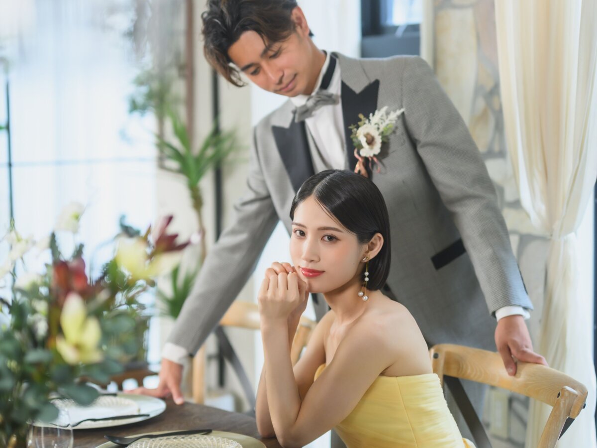 テーブルに頬杖をつくイエローのカラードレスの花嫁と、後ろから寄り添うグレーのタキシードの新郎。カフェのような空間で撮るおしゃれな前撮り