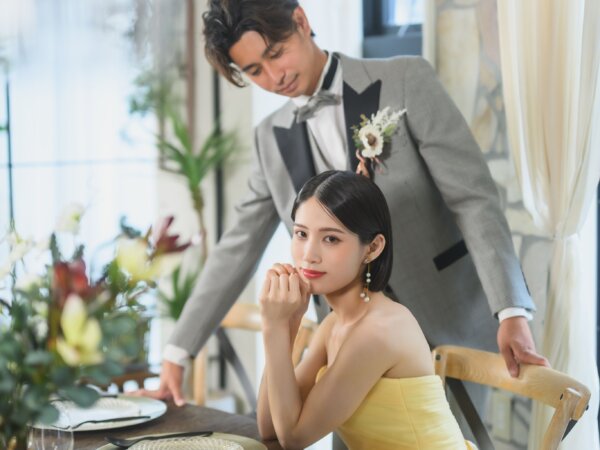 テーブルに頬杖をつくイエローのカラードレスの花嫁と、後ろから寄り添うグレーのタキシードの新郎。カフェのような空間で撮るおしゃれな前撮り