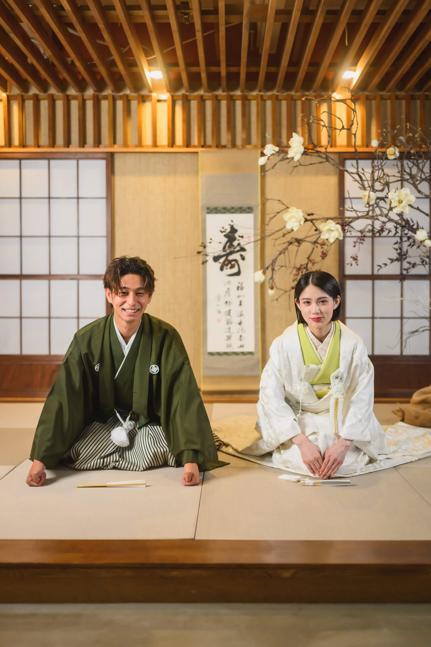 '寿'の掛け軸と白木蓮の生け花を背に、畳の上へ並んで正座する白無垢の花嫁と紋付袴の新郎。凛とした佇まいの和装フォトウェディング