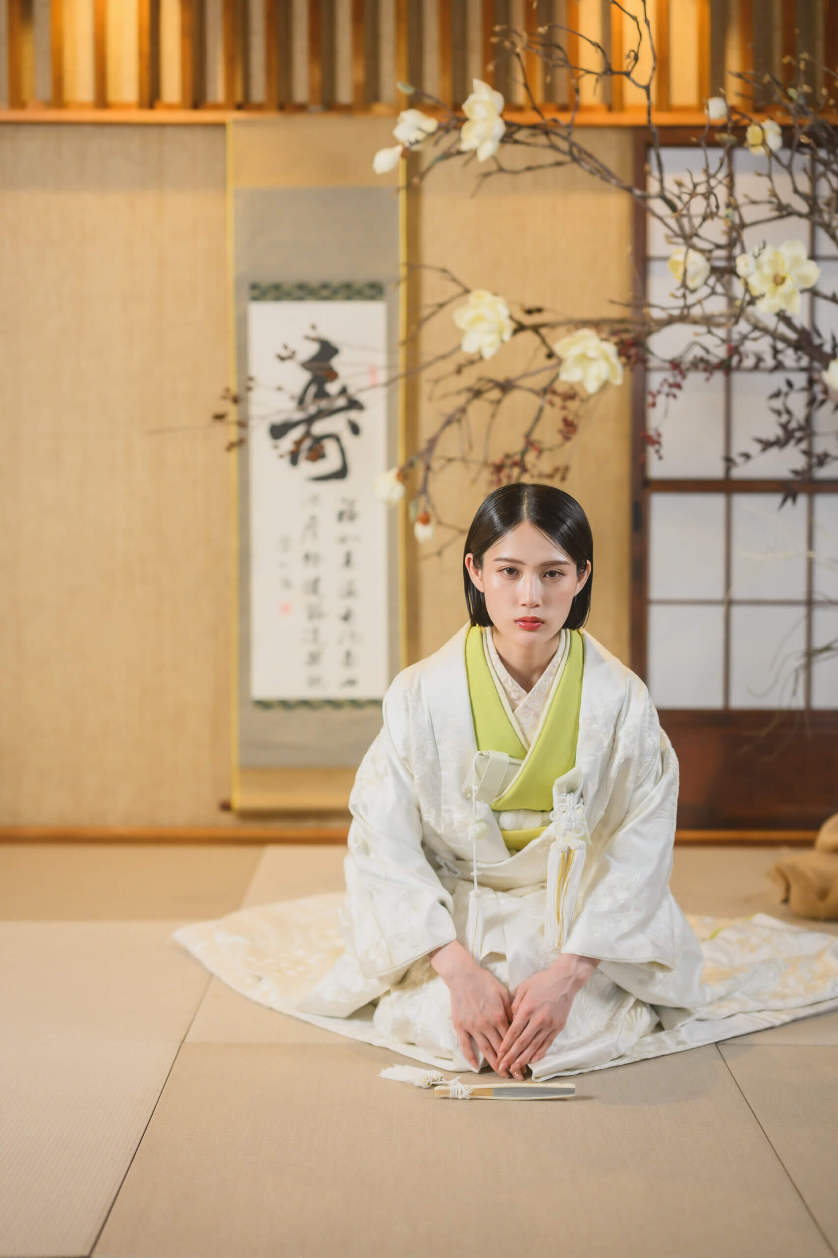 畳の間に正座して正面を見つめる白無垢の花嫁。黄緑の掛下と白木蓮の生け花が引き立つ和装前撮りのポートレート