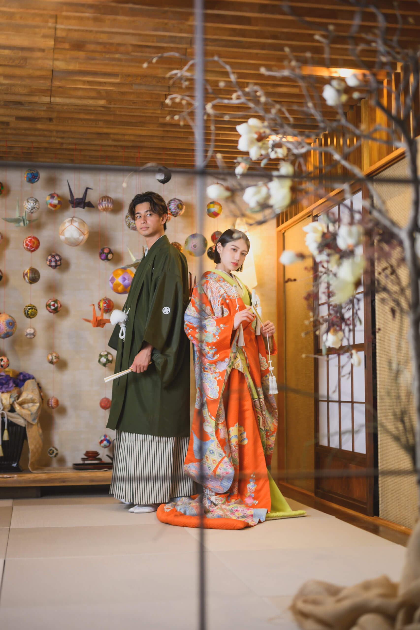 手毬の壁飾りの前で背中合わせに立つ、オレンジの色打掛の花嫁と深緑の紋付袴の新郎。扇子を手にした和装前撮りのポーズ
