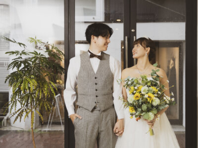 ガラス扉の前で手をつないで見つめ合う、グレーのベストの新郎とひまわりのブーケを持つウェディングドレスの花嫁。名古屋で撮る結婚写真