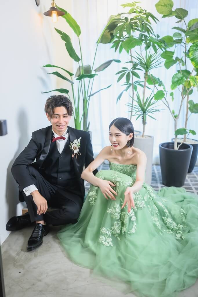 観葉植物に囲まれた床に腰を下ろし、笑い合うグリーンのカラードレスの花嫁と黒タキシードの新郎。名古屋・栄のスタジオで撮るフォトウェディング