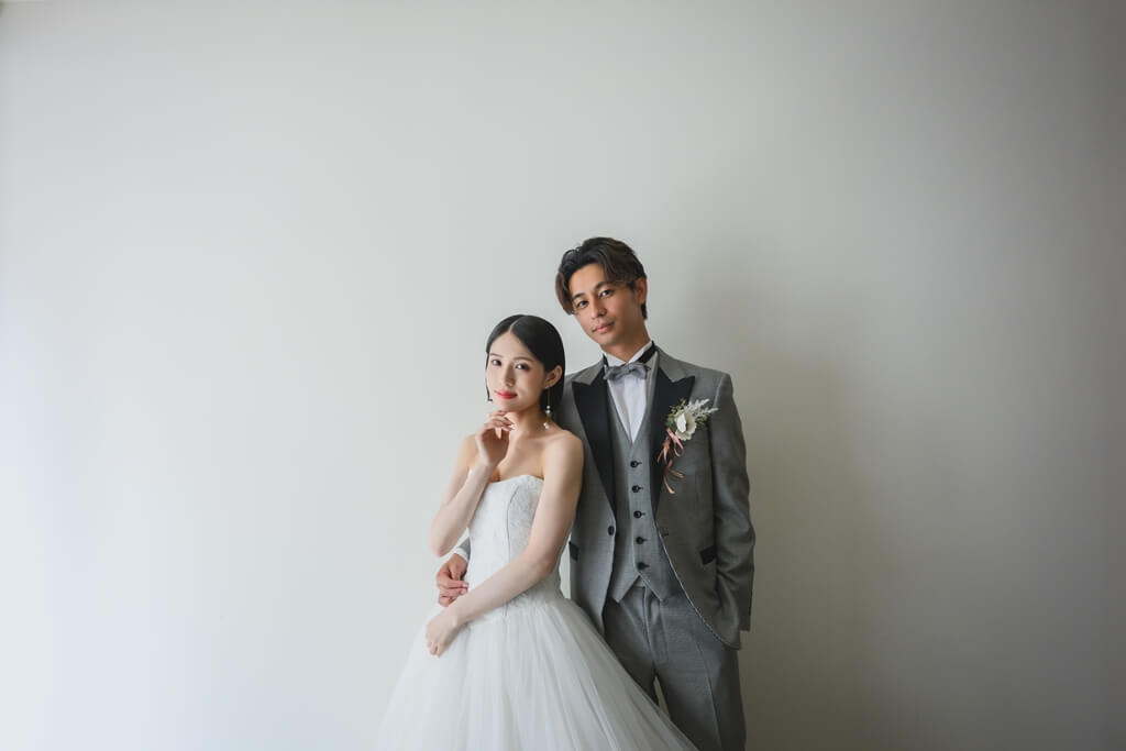真っ白な壁を背景に、頬に手を添えて寄り添うウェディングドレスの花嫁とグレーのタキシードの新郎。名古屋・栄のスタジオで撮る洋装フォトウェディング