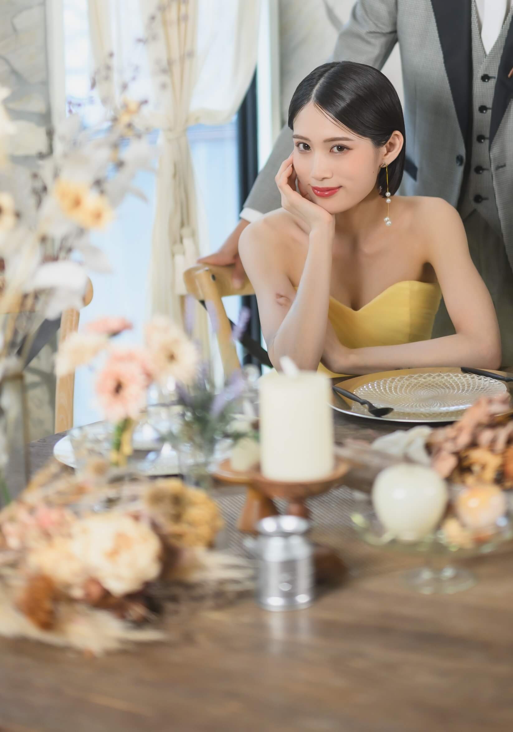 ドライフラワーを飾ったテーブルで頬杖をつくイエローのカラードレスの花嫁と、傍らに立つグレータキシードの新郎。結婚写真のワンシーン