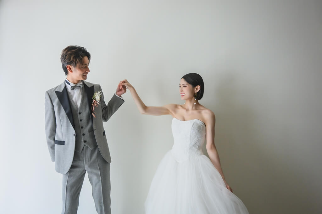 白い壁の前で花嫁の手を取りエスコートする新郎。チュールのウェディングドレスとグレータキシードで残す前撮りのワンシーン