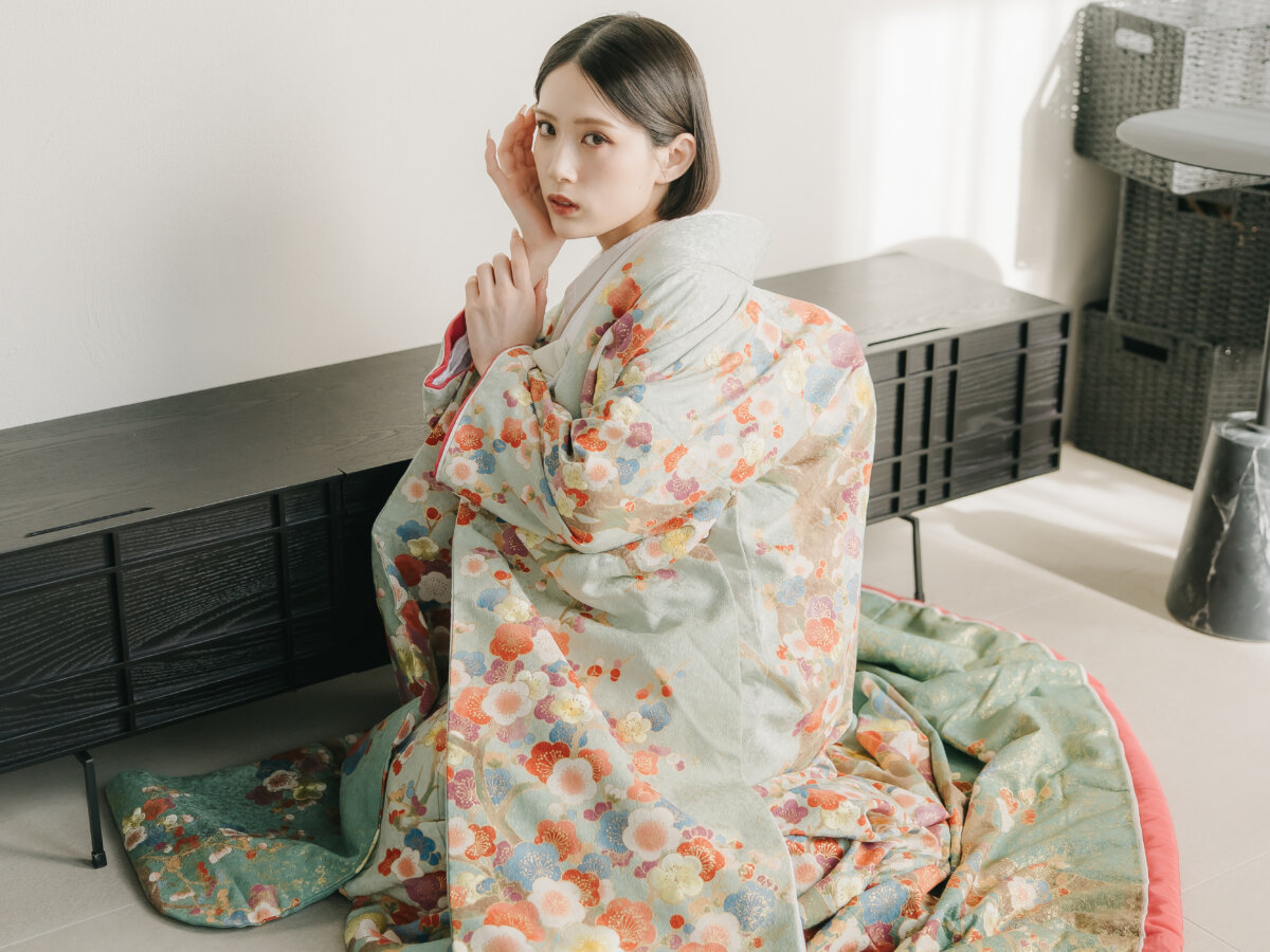 白い壁のスタジオで床に座り、頬に手を添えて振り向く、若草色に花模様の色打掛の花嫁。モダンな和装前撮りのポーズ