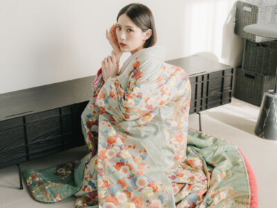 白い壁のスタジオで床に座り、頬に手を添えて振り向く、若草色に花模様の色打掛の花嫁。モダンな和装前撮りのポーズ
