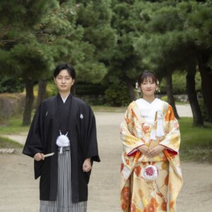 松の木立の小道に並んで立つ、黒紋付袴の新郎と金色に菊の柄の色打掛の花嫁。白鳥庭園で撮る色打掛の和装前撮り