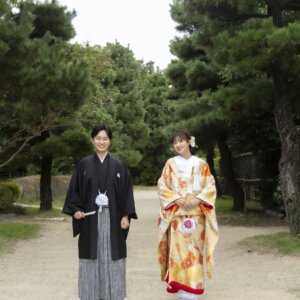 松に囲まれた白鳥庭園の園路で、末広を手に並ぶ縞袴の新郎と金地に赤い花柄の色打掛の花嫁。秋の和装ロケーションフォト