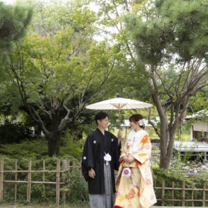 松の木の下で白い和傘をさし、竹の柵のそばに寄り添う黒紋付袴の新郎と色打掛の花嫁。白鳥庭園の和装前撮り